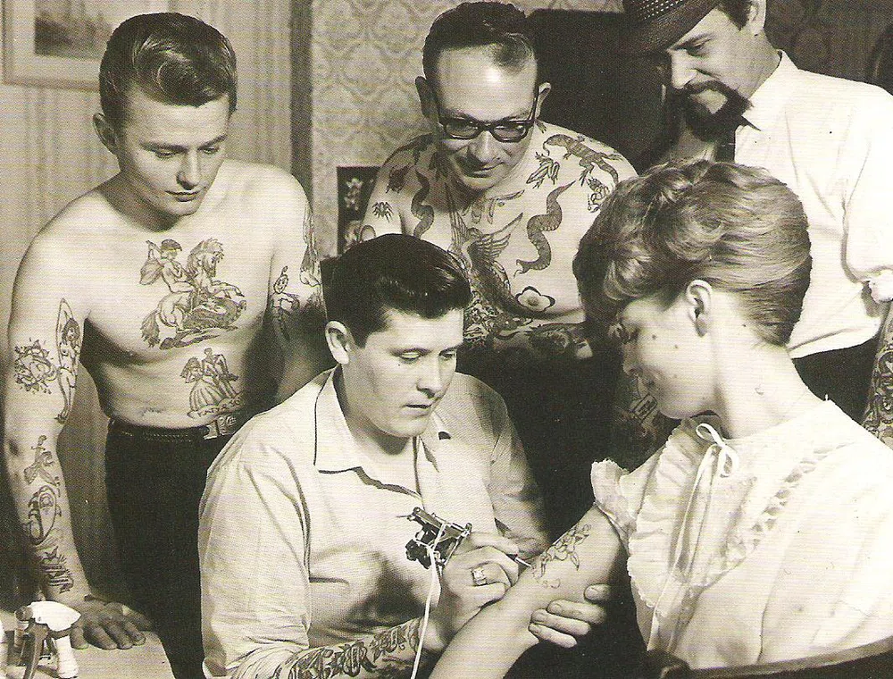 fotografía en blanco y negro de principios del siglo XX. Realizando un tatuaje en el brazo de una mujer delante de un grupo de personas que muestran orgullosos sus tatuajes.