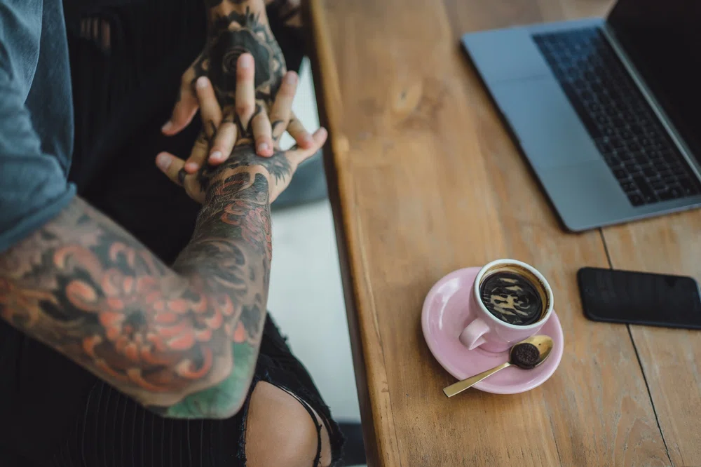 Tattooed hands leaning on a table with a coffee and a laptop